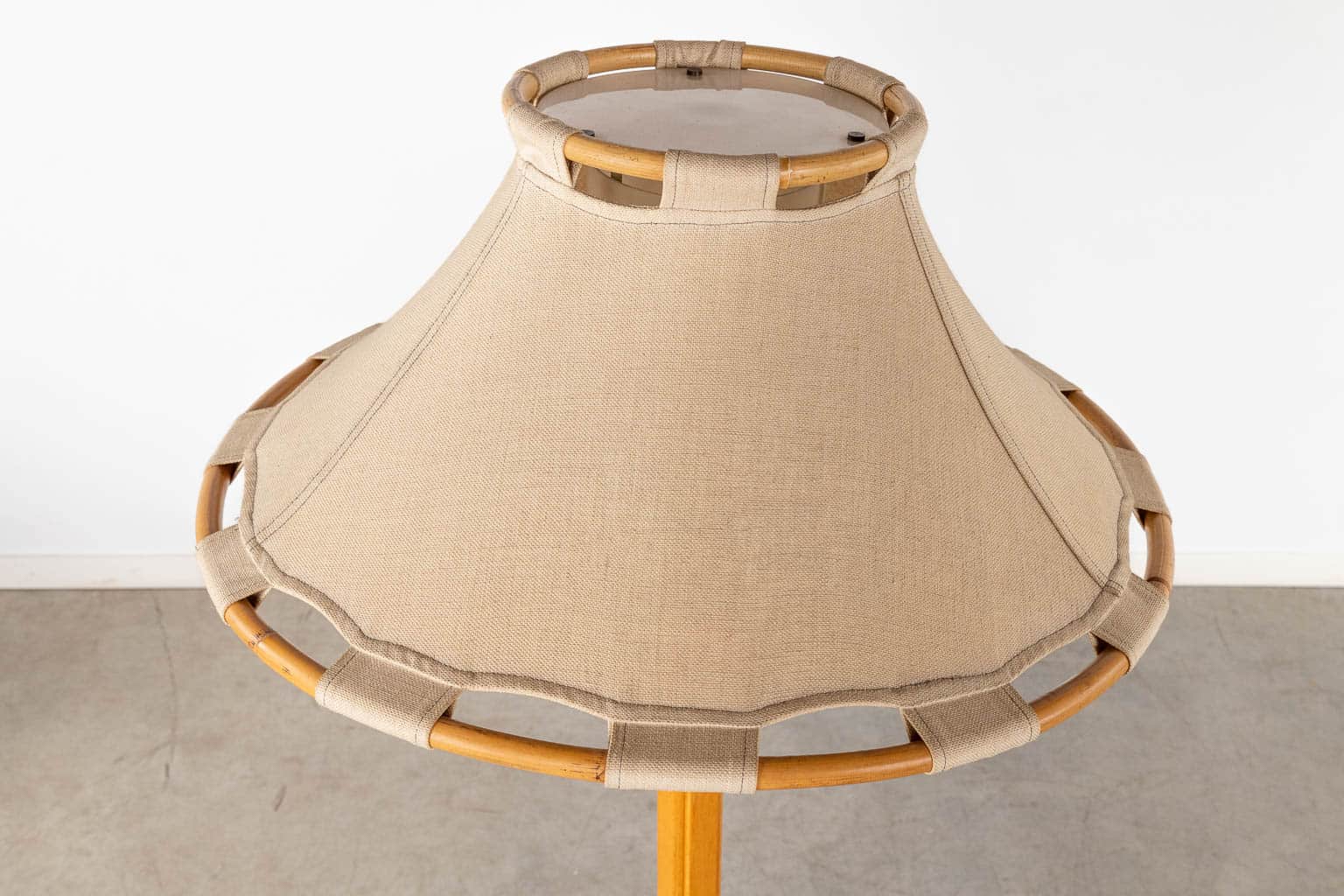 Een close-up van een beige stoffen lampenkap met een houten frame en decoratieve lussen rond de rand, tegen een lichtgekleurde muur en grijze vloer.