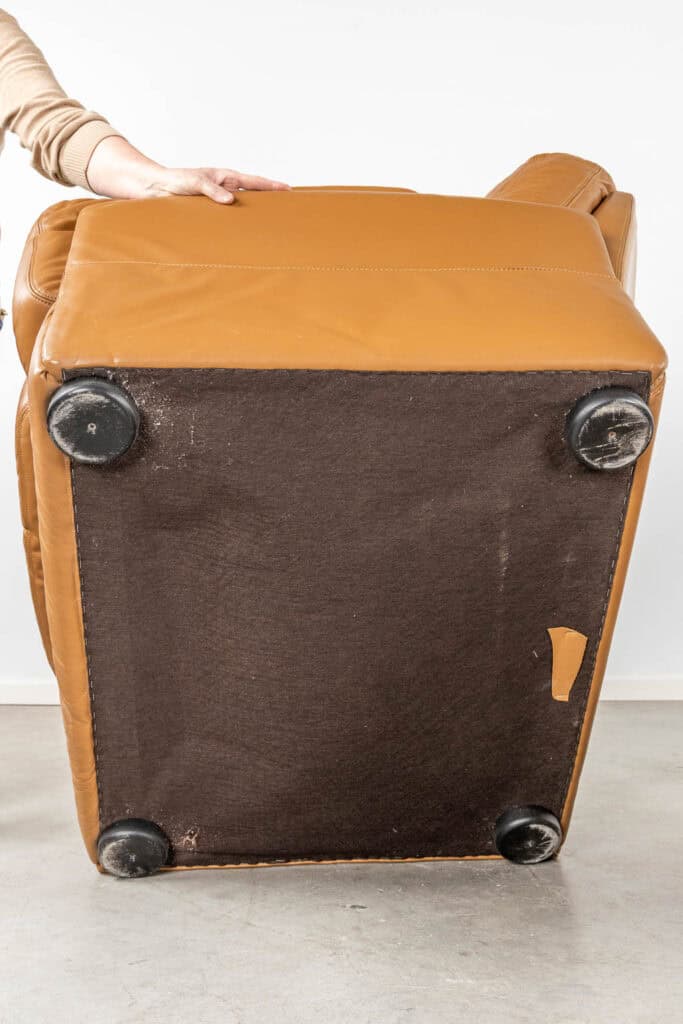 Een persoon in een beige hoes houdt een beige leren fauteuil omhoog, waarvan de onderkant met vier zwarte plastic poten en een donkere stoffen bekleding zichtbaar is. Een klein stukje beige stof is losgelaten van de stof.