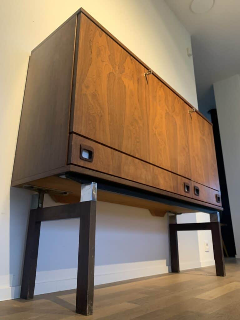Een houten kast in mid-century modern-stijl met vierkante handgrepen en metalen poten staat tegen een witte muur op een houten vloer, bekeken vanuit een laag perspectief.