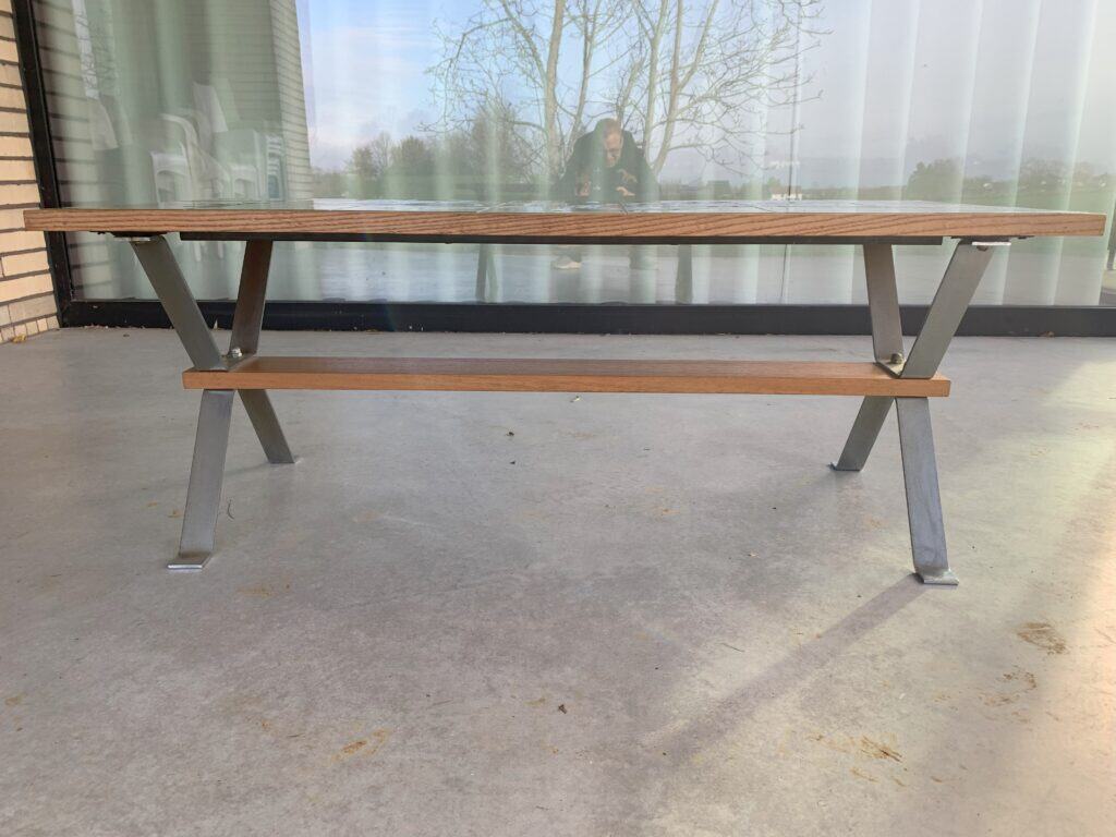 Een houten bank met metalen poten staat op een betonnen ondergrond voor een glazen wand. In het glas achter de bank zijn de weerspiegeling van een persoon en een boom te zien.