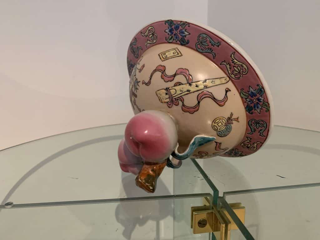 Een decoratieve keramische kom met roze en bloemmotieven, ondersteboven op een glazen plankje. De kom heeft een ronde, gebeeldhouwde roze en blauwe voet in de vorm van een dier.