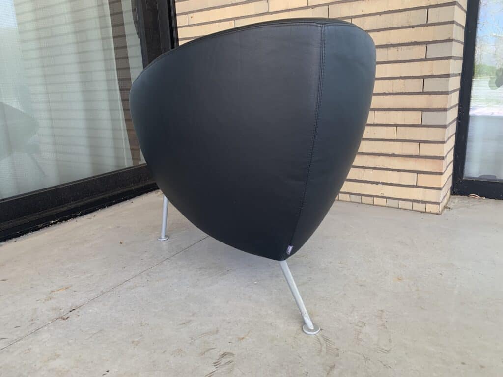 Een moderne, zwarte, leren stoel met een ronde rugleuning en drie dunne metalen poten staat op een betonnen terras bij een bakstenen muur en een glazen schuifdeur.