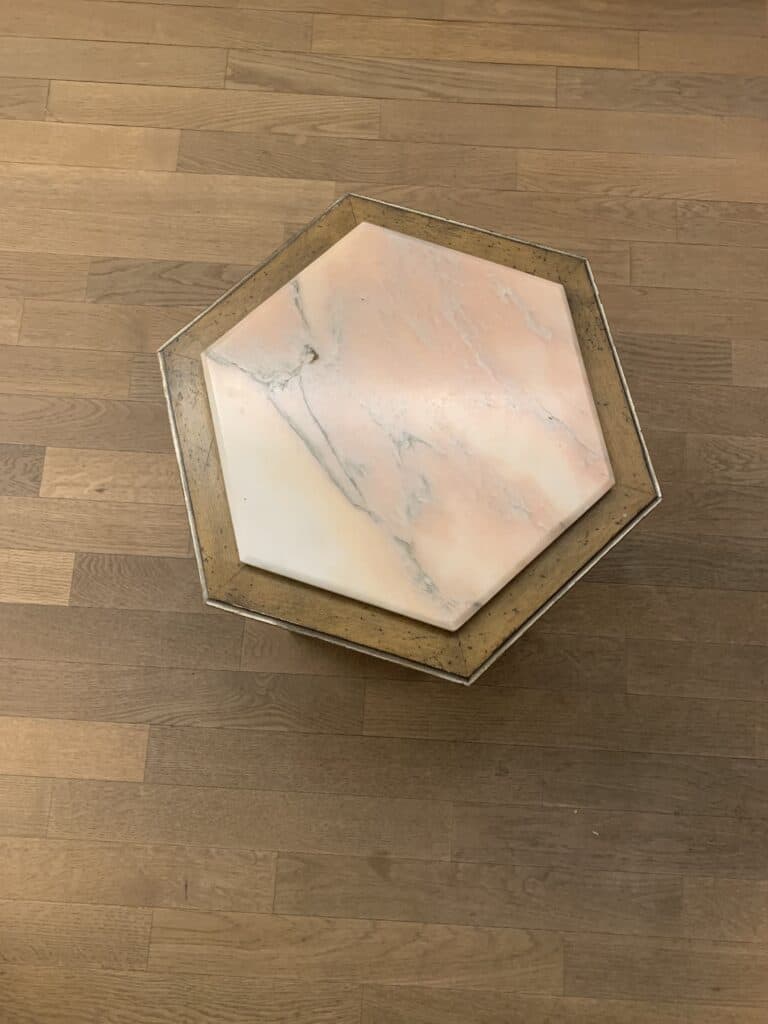 Een zeshoekige tafel met een marmeren blad met lichtroze en grijze nerven, geplaatst in een goudkleurig frame, op een houten vloer met een middelbruine afwerking.