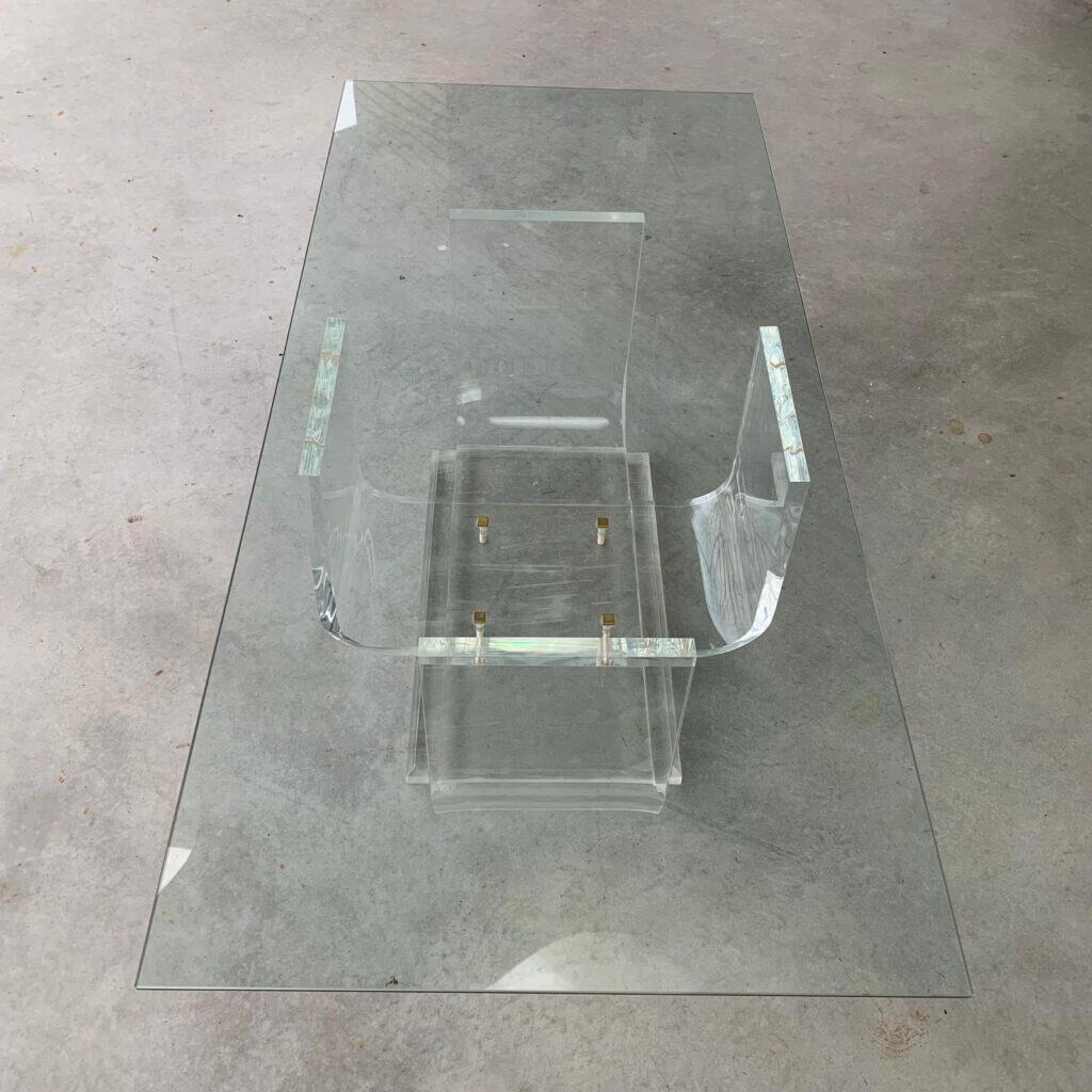 Een salontafel van helder glas met een sculpturaal acryl onderstel, geplaatst op een lichtgrijze betonnen vloer. Het ontwerp is modern en minimalistisch, met strakke, transparante lijnen en metalen accenten op het onderstel.