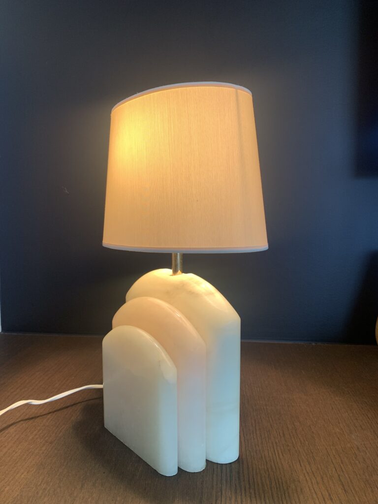 Een moderne tafellamp met een beige kap en een sculpturale witte marmeren voet met drie ronde, op elkaar gestapelde lagen, staat op een donker houten oppervlak tegen een donkere muur.