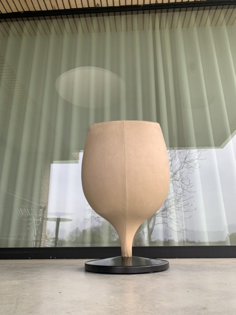 Een grote, beige, wijnglasvormige stoel met een ronde kom en een smalle basis staat op een zwart rond platform voor een glazen wand met verticale gordijnen.