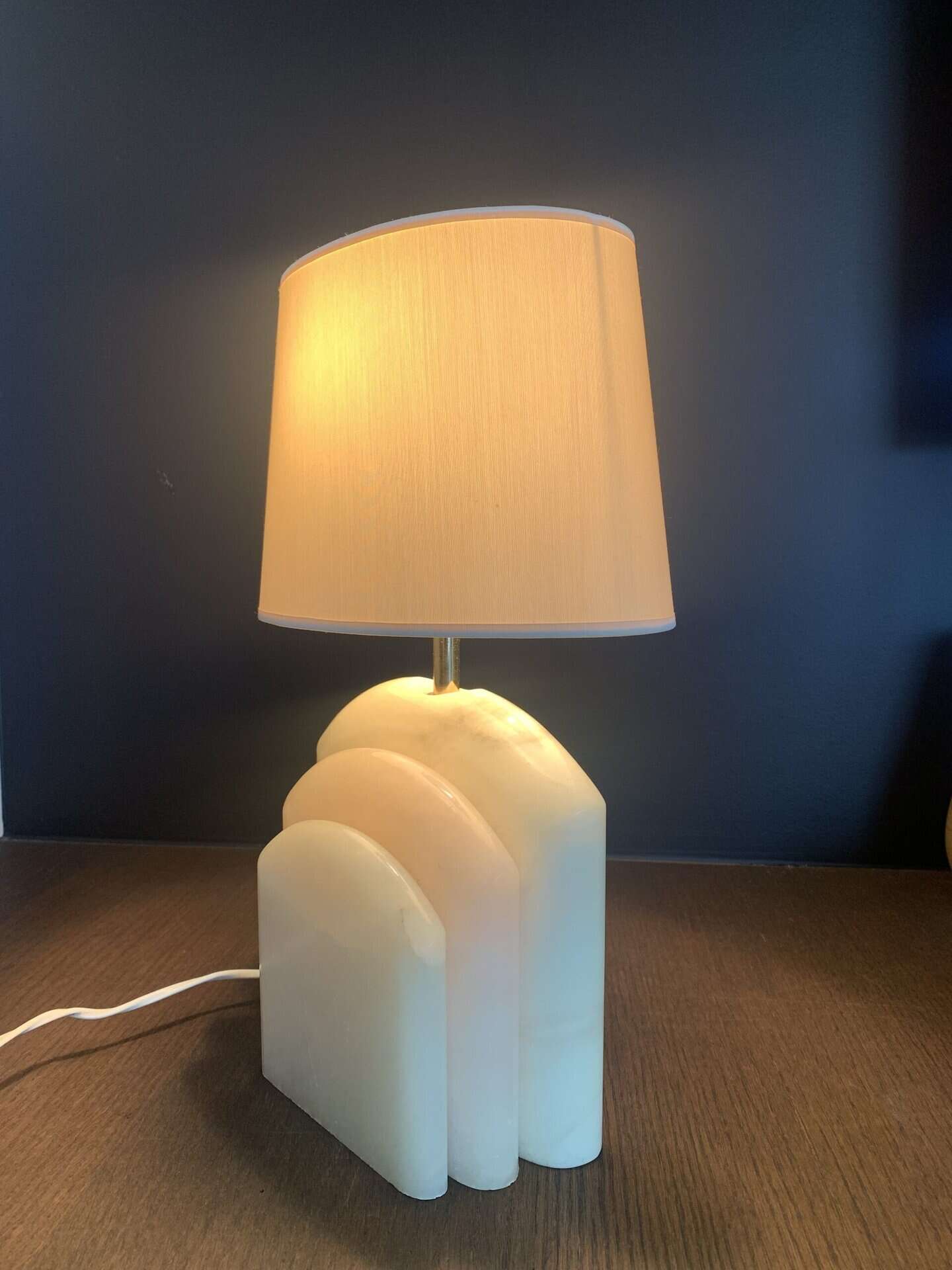 Een moderne tafellamp met een beige kap en een sculpturale witte marmeren voet met drie ronde, op elkaar gestapelde lagen, staat op een donker houten oppervlak tegen een donkere muur.