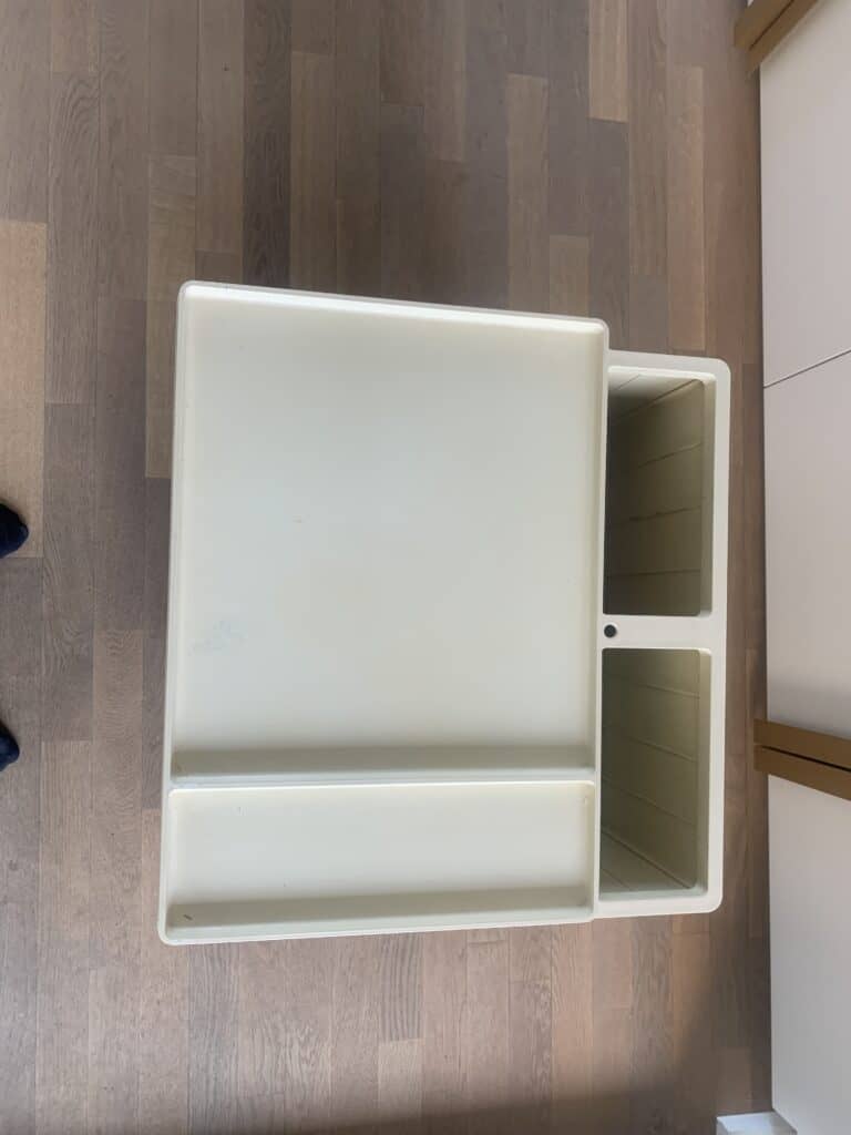 Een wit, rechthoekig, kunststof bureau met twee open vakken aan één kant, geplaatst op een houten vloer. Het bureau is van bovenaf te zien, met de verdeelde opbergruimtes.