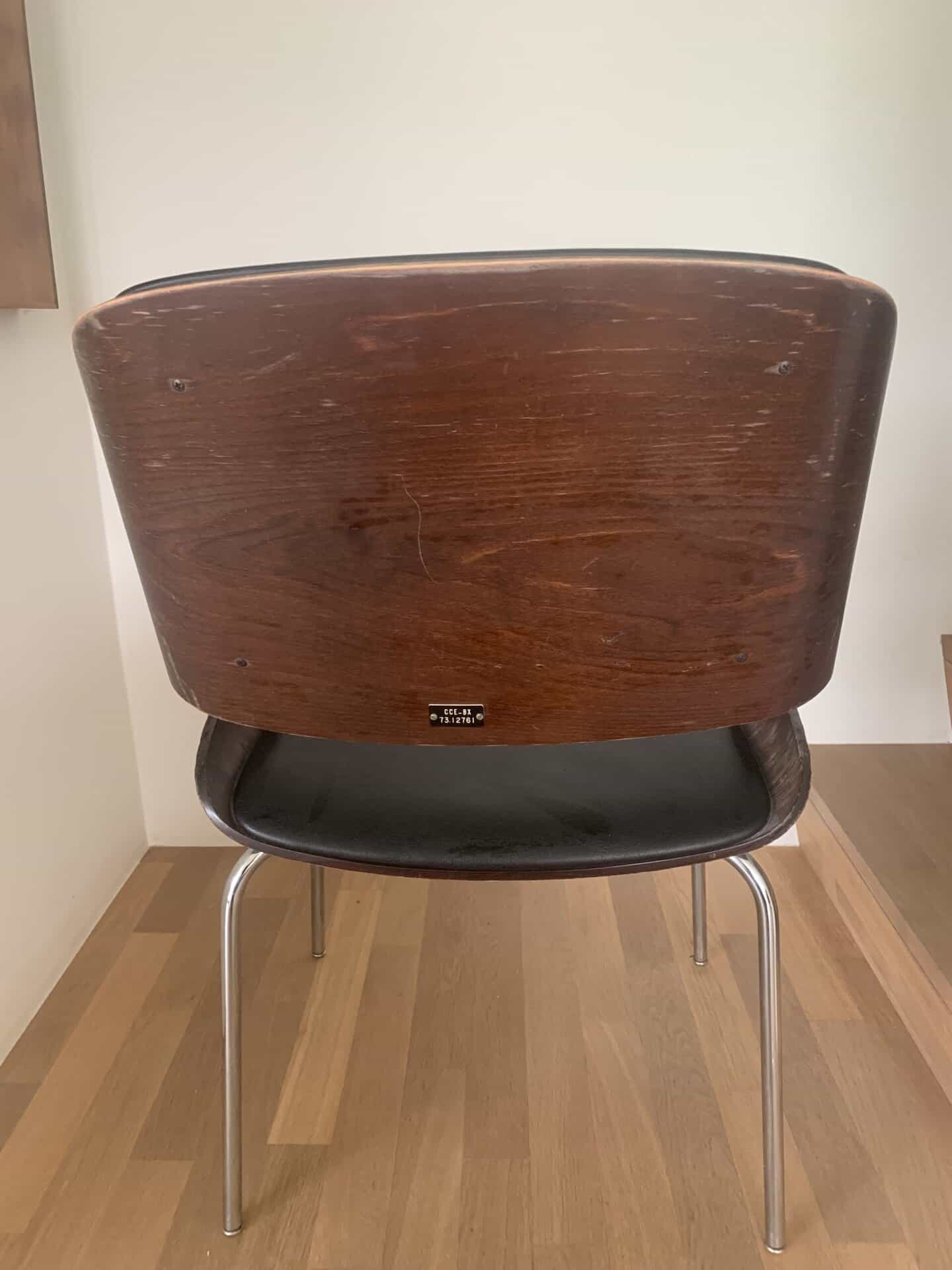 Een houten stoel met een gebogen rugleuning en metalen poten staat op een lichtgekleurde houten vloer, van de camera af gericht. Aan de rugleuning is een klein zwart label bevestigd.