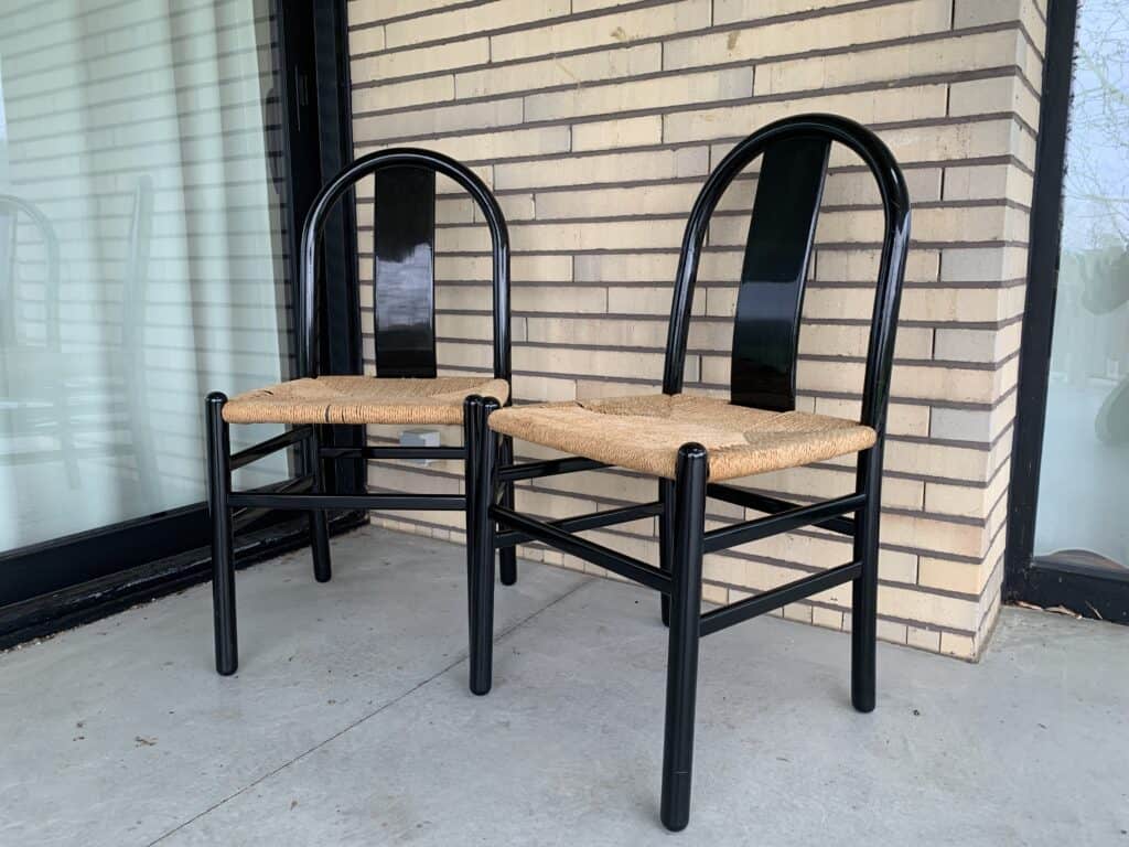 Twee zwarte houten stoelen met gebogen rugleuningen en gevlochten rieten zittingen staan naast elkaar op een betonnen vloer, voor een bakstenen muur en een groot raam.