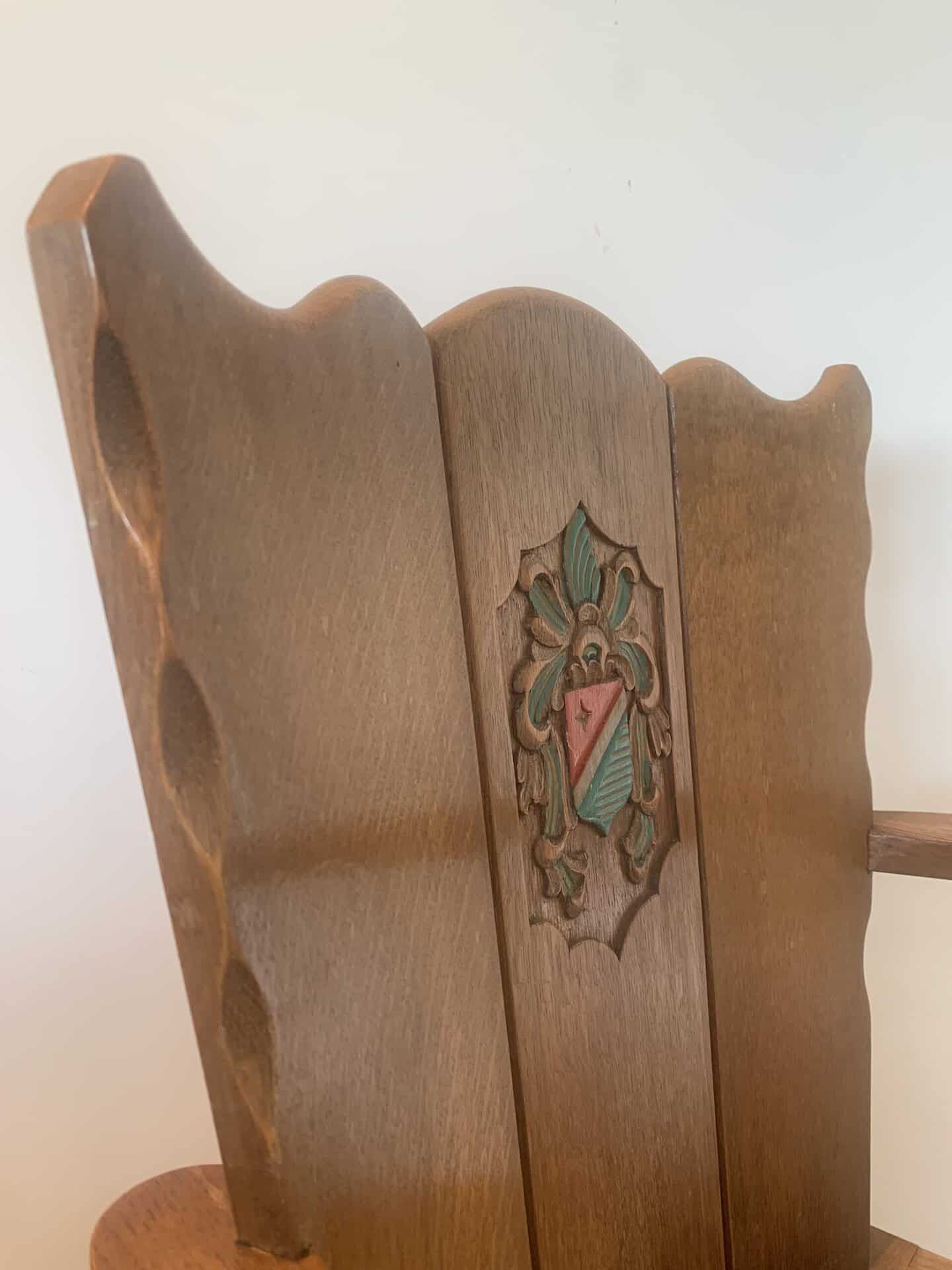 Een houten stoel met een hoge, sierlijke rugleuning met een gebeeldhouwd en beschilderd wapen met een rode driehoek en groene details, tegen een eenvoudige, lichte achtergrond.