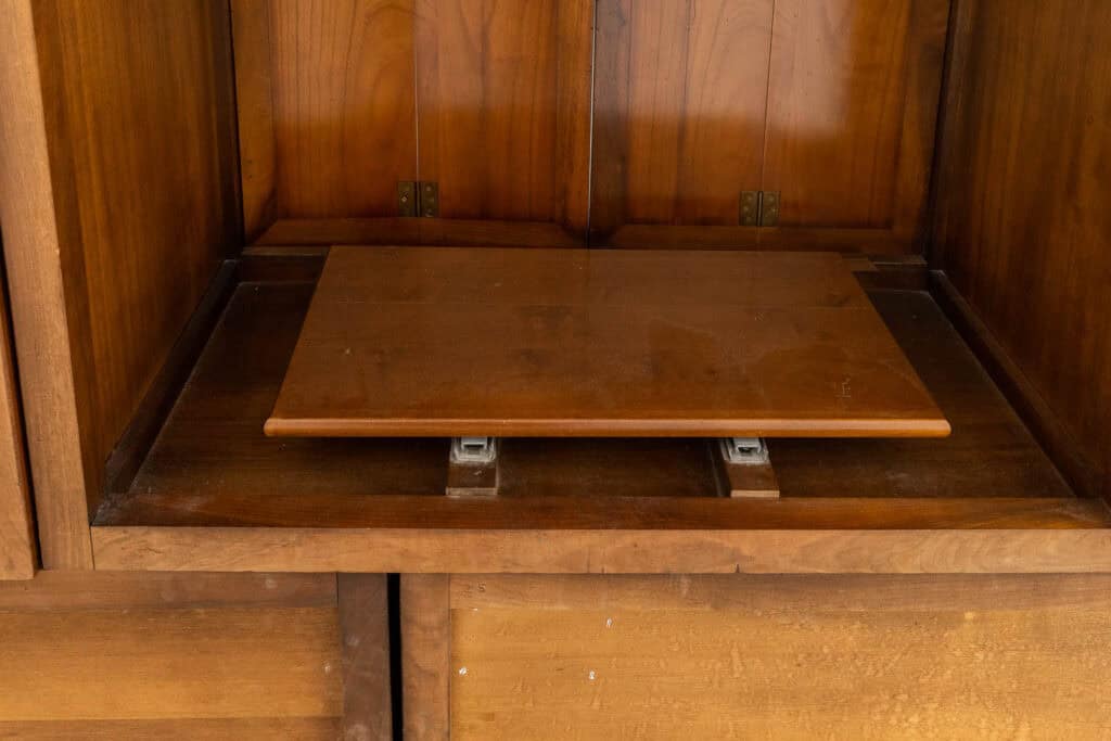 Een close-up van een houten kast met een uitschuifbare plank die gedeeltelijk is uitgeklapt, waardoor de metalen schuifmechanismen eronder zichtbaar zijn. Het hout heeft een warme, natuurlijke afwerking.