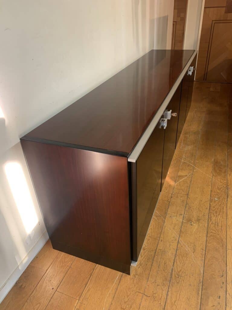 Een donkerhouten dressoir met een glanzende afwerking en zilveren handgrepen staat tegen een lichte muur op een houten vloer, verlicht door zonlicht van links.