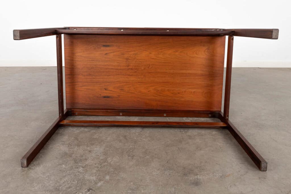 Een houten tafel staat op zijn kop, waarbij de onderkant en de poten zichtbaar zijn. De tafel staat op een betonnen vloer met een eenvoudige witte muur op de achtergrond.