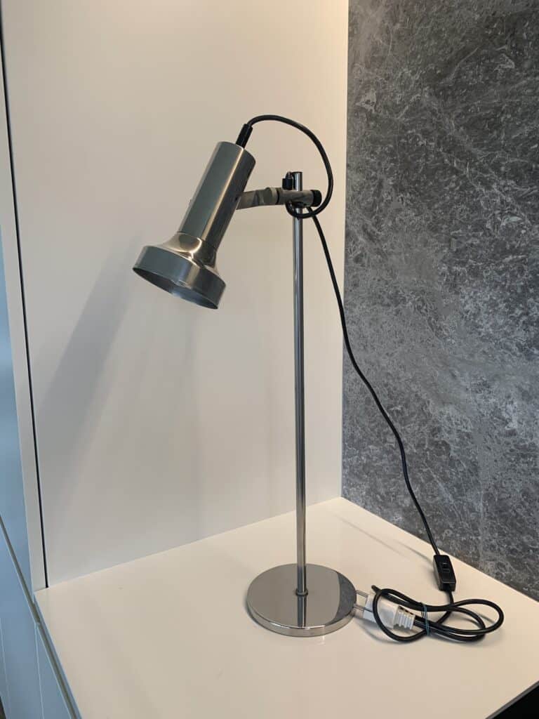 Een moderne, zilveren bureaulamp met een cilindrische kap staat op een wit oppervlak naast een grijs gemarmerde muur. Het zwarte snoer van de lamp is zichtbaar en aangesloten, met een schakelaar op het snoer.