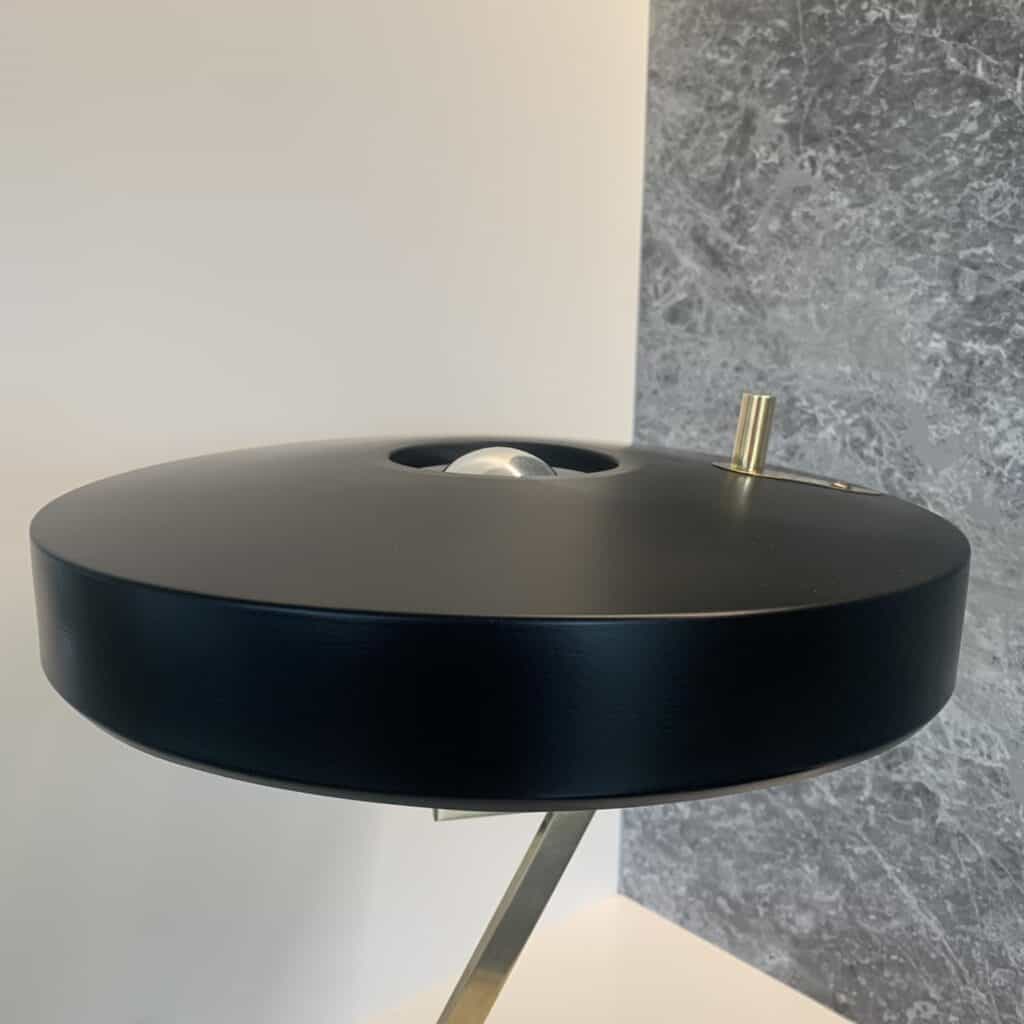 Een moderne, zwarte bureaulamp met een ronde, platte lampenkap en metalen accenten staat voor een witte muur en een grijs oppervlak met structuur.
