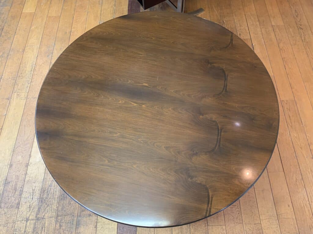 Van bovenaf is een ronde houten tafel met een donkere afwerking te zien, met zichtbare houtnerfpatronen en drie gebogen lijnen aan de rechterkant. De tafel staat op een lichte houten vloer.