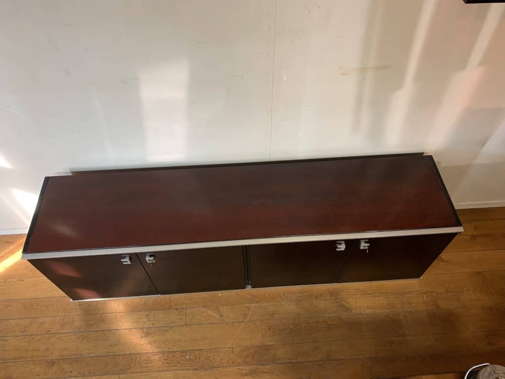 Een donkere houten kast met zilveren handgrepen staat tegen een witte muur op een lichtbruine houten vloer, van bovenaf gezien.