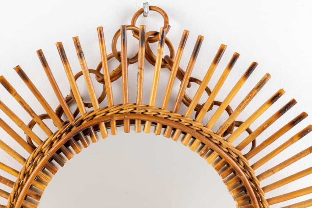 Een close-up van het bovenste gedeelte van een ronde rieten spiegel met een decoratief, zonnestralenmotief, gemaakt van gevlochten en gebogen rieten stroken tegen een witte muur.