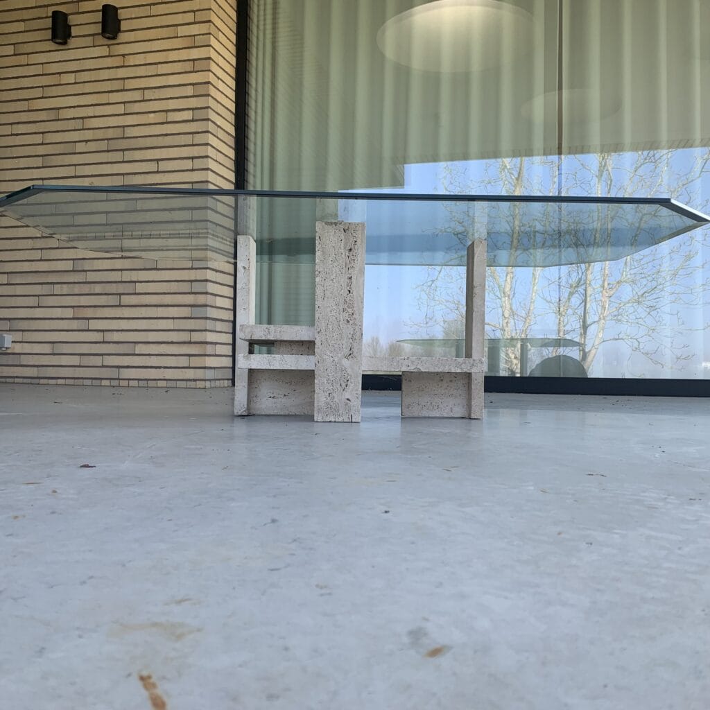 Een moderne tafel met glazen blad en geometrische stenen poten staat op een gladde terrasvloer voor grote glazen deuren en beige bakstenen muren. In het raam zijn de weerspiegelingen van bomen zichtbaar.