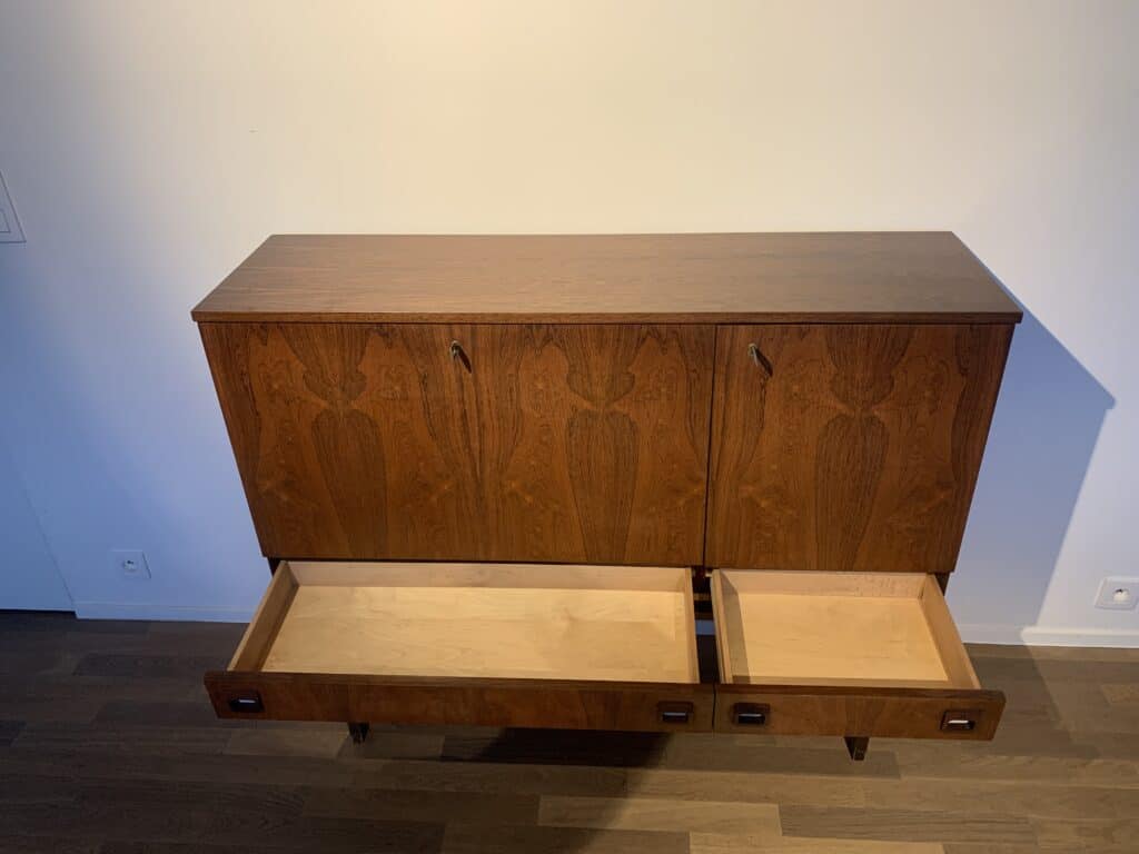 Een houten kast met drie gesloten bovendeuren en twee onderste laden, waarvan er één open is, staat tegen een witte muur op een hardhouten vloer.