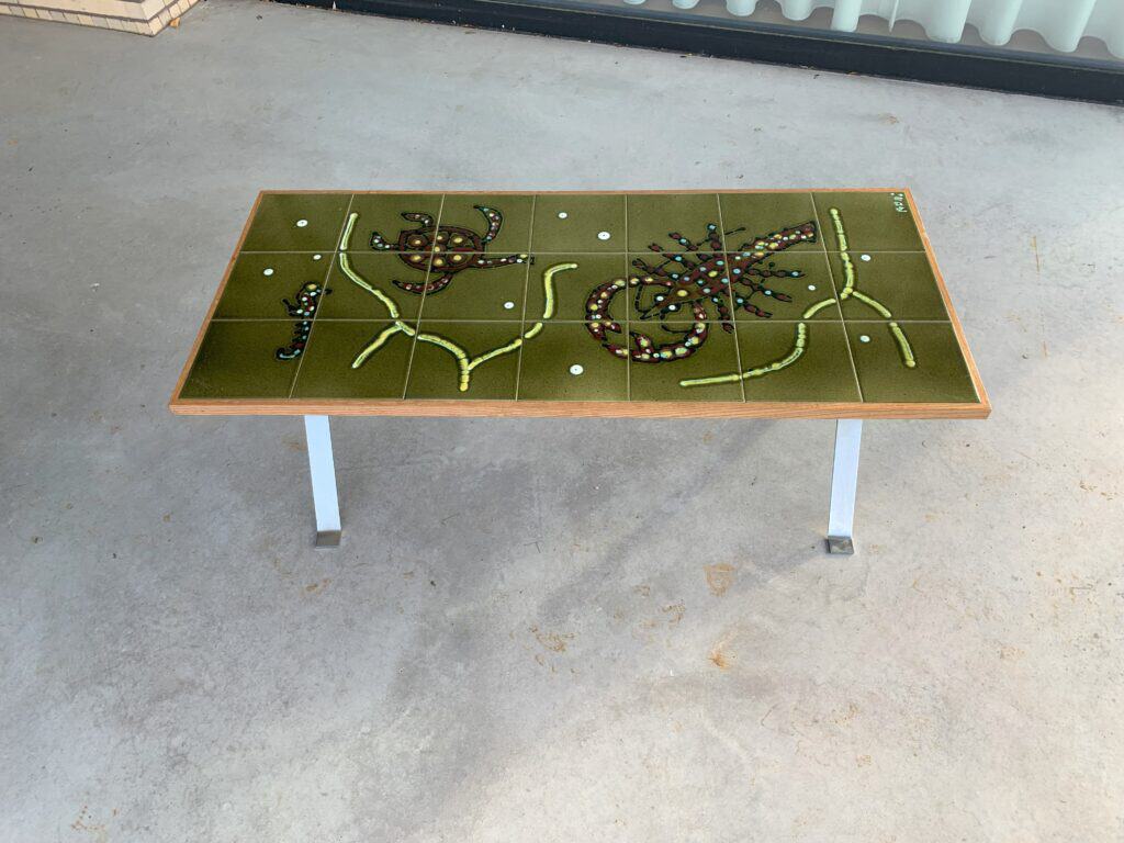 Een rechthoekige tafel met een groen oppervlak heeft een abstract ontwerp van kralen en kleine voorwerpen, die lijken op planten of dieren. De tafel heeft metalen poten en staat op een betonnen vloer.