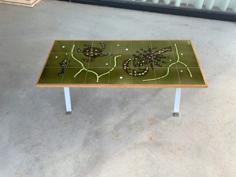Een rechthoekige tafel met een groen oppervlak heeft een abstract ontwerp van kralen en kleine voorwerpen, die lijken op planten of dieren. De tafel heeft metalen poten en staat op een betonnen vloer.