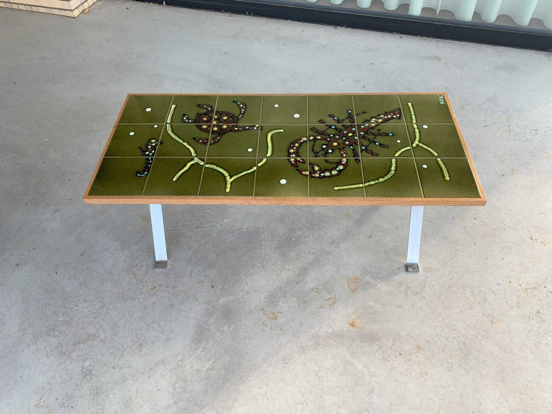 Een rechthoekige tafel met een groen oppervlak heeft een abstract ontwerp van kralen en kleine voorwerpen, die lijken op planten of dieren. De tafel heeft metalen poten en staat op een betonnen vloer.