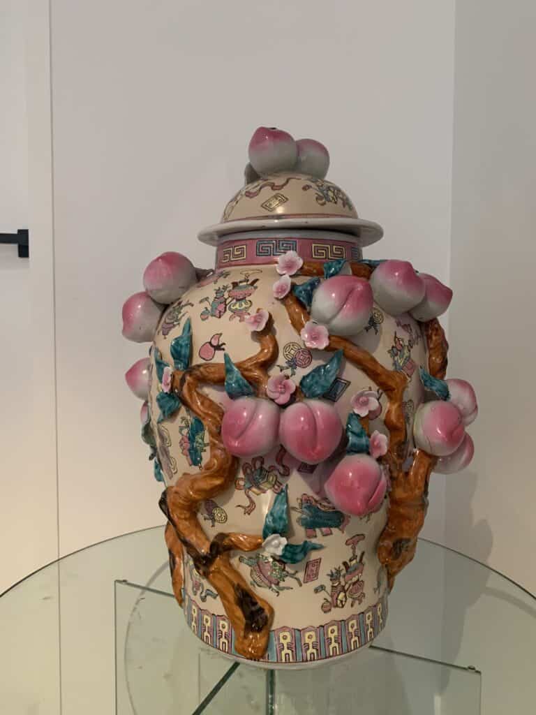 Een decoratieve keramische vaas met deksel, versierd met roze perziken, bruine takken, groene bladeren en kleurrijke vogels, zittend op een glazen oppervlak tegen een witte achtergrond.