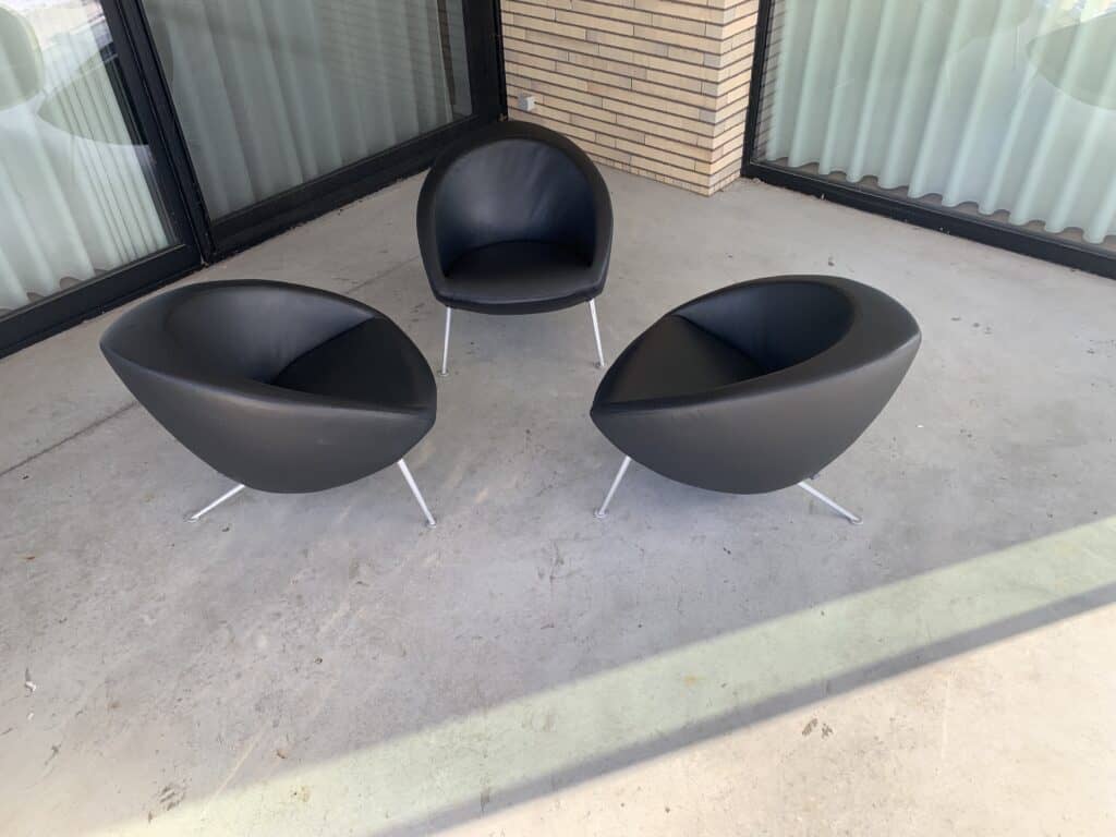 Drie moderne, zwarte fauteuils met gebogen rugleuningen staan in een halve cirkel op een betonnen terras naast een gebouw met bakstenen muren en grote glazen ramen.