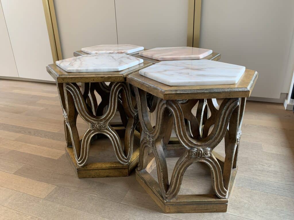 Vier zeshoekige tafels met sierlijke houten onderstellen en witte marmeren bladen staan dicht bij elkaar op een houten vloer, voor lichtgekleurde kasten.