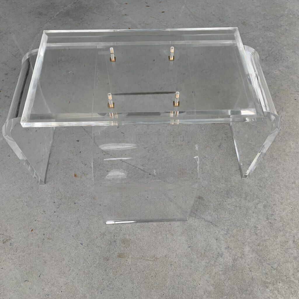Een transparante tafel van acryl met een rechthoekig blad, gebogen zijkanten en goudkleurige bevestigingsmiddelen, geplaatst op een lichtgrijze betonnen vloer.