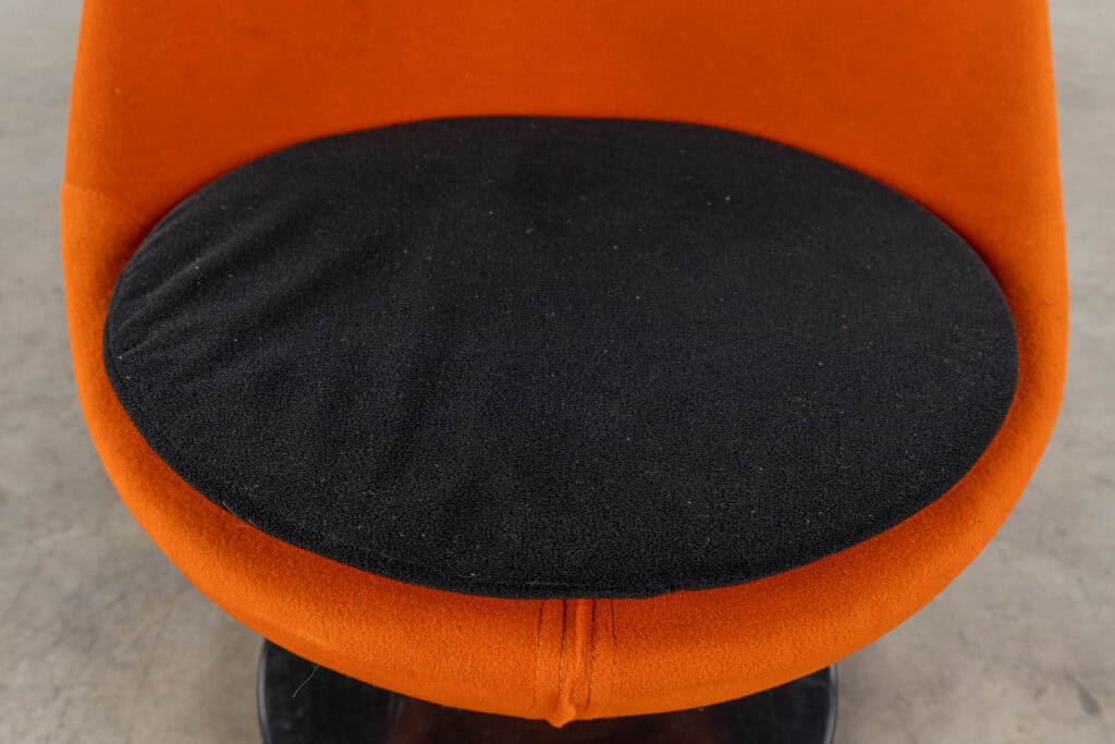 Een close-up van een oranje stoel met een rond, zwart zitkussen, tegen een neutrale, lichtgrijze achtergrond.