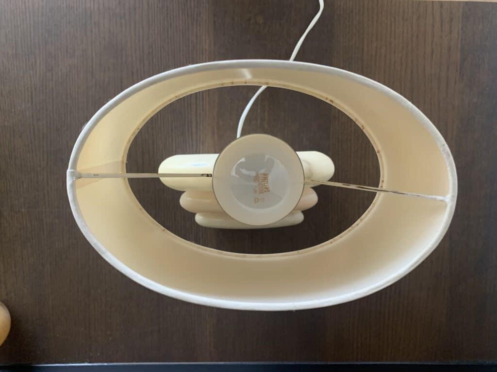 Bovenaanzicht van een tafellamp met een ovale, beige lampenkap en een witte gloeilamp, geplaatst op een donker houten oppervlak. Het witte snoer van de lamp steekt omhoog uit het frame.