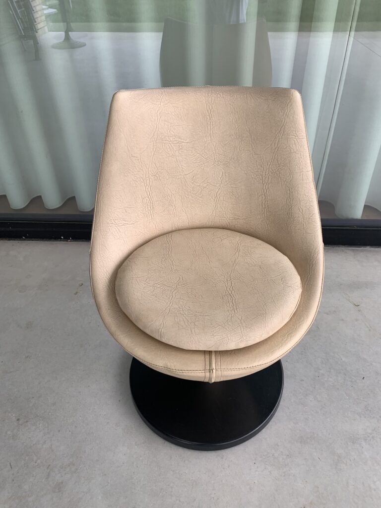 Een beige, moderne draaistoel met een rond zitkussen en een zwart onderstel staat op een lichte vloer voor een glazen deur met witte gordijnen.