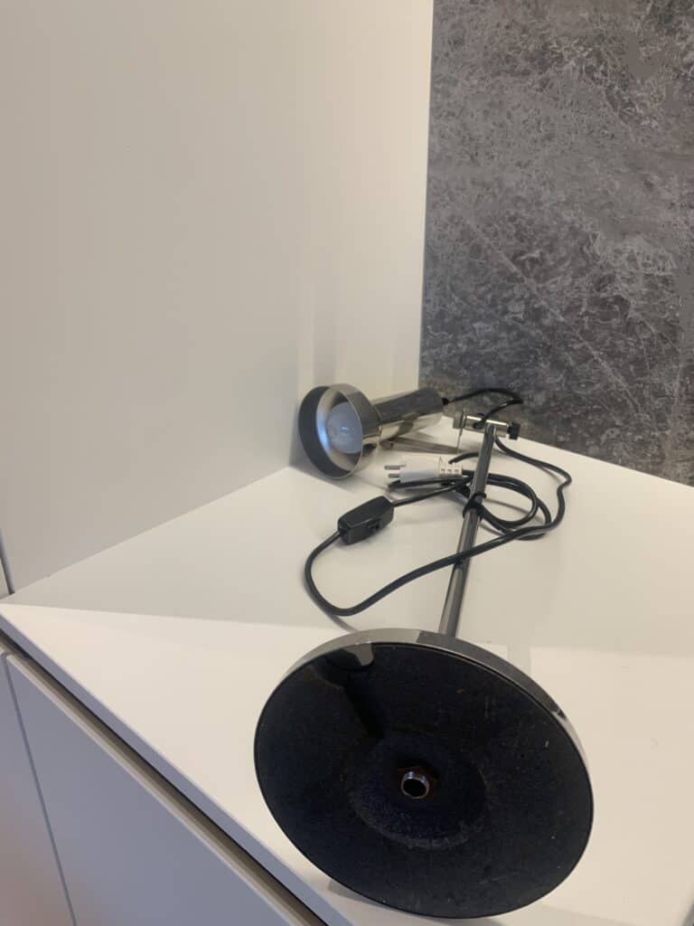 Een zwarte bureaulamp ligt gedemonteerd op een wit aanrecht, met de voet, kap, het netsnoer en de schakelaar zichtbaar. De achtergrond bestaat uit een witte muur en een grijs getextureerd oppervlak.