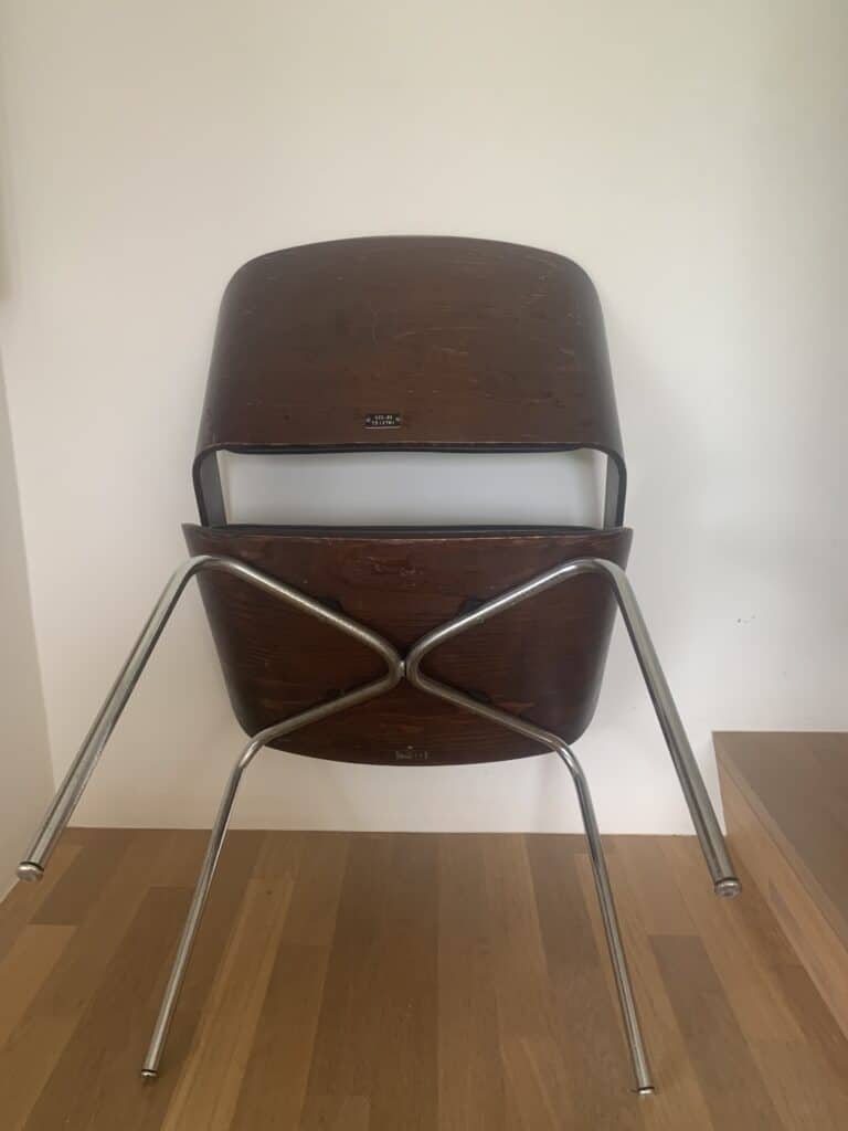Twee houten stoelen met metalen poten staan ondersteboven opgestapeld tegen een witte muur en naast een houten trap. De poten van de bovenste stoel rusten op de vloer.