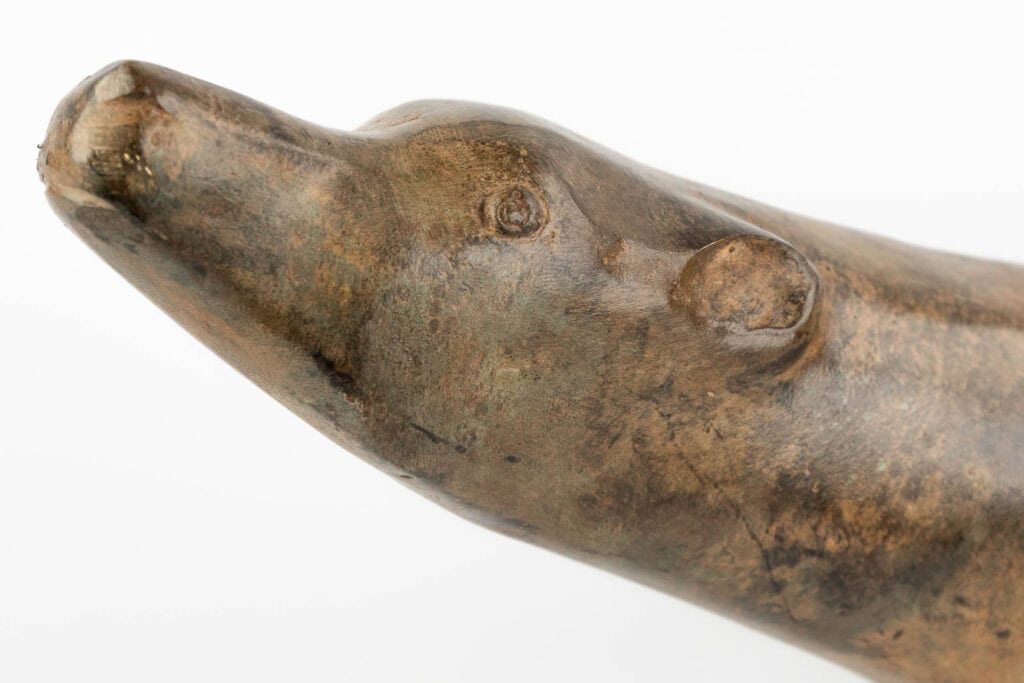 Een close-up van een bronzen of bruin gebeeldhouwde dierenkop, mogelijk een beer, afgebeeld tegen een witte achtergrond. Het beeld heeft een glad, licht getextureerd oppervlak met eenvoudige, gestileerde kenmerken.