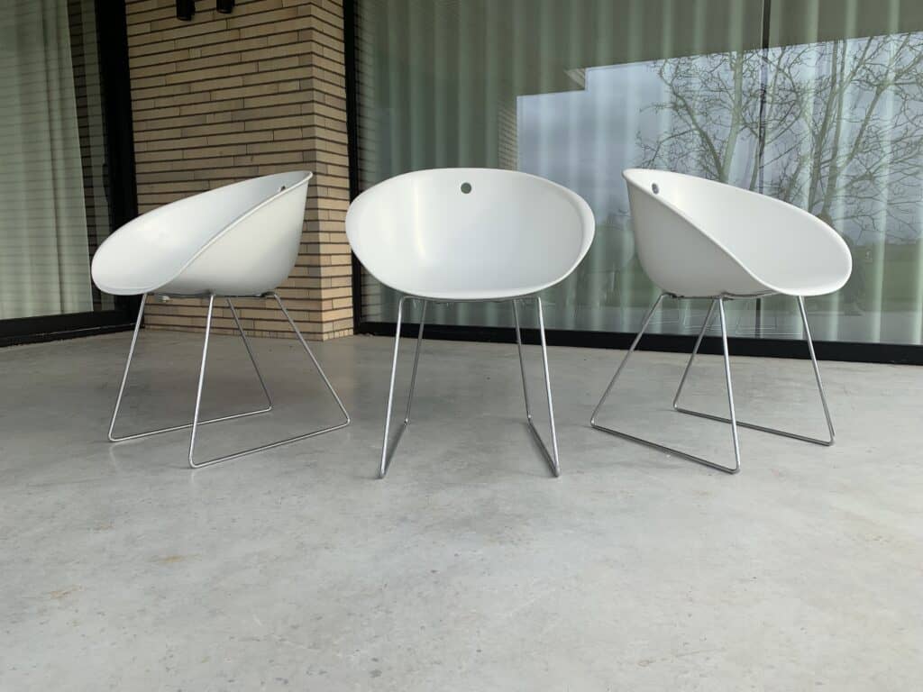 Drie moderne witte stoelen met metalen poten staan op een betonnen terras voor een glazen raam en een gebouw met lichte bakstenen muren. In het raam is een weerspiegeling van bomen te zien.