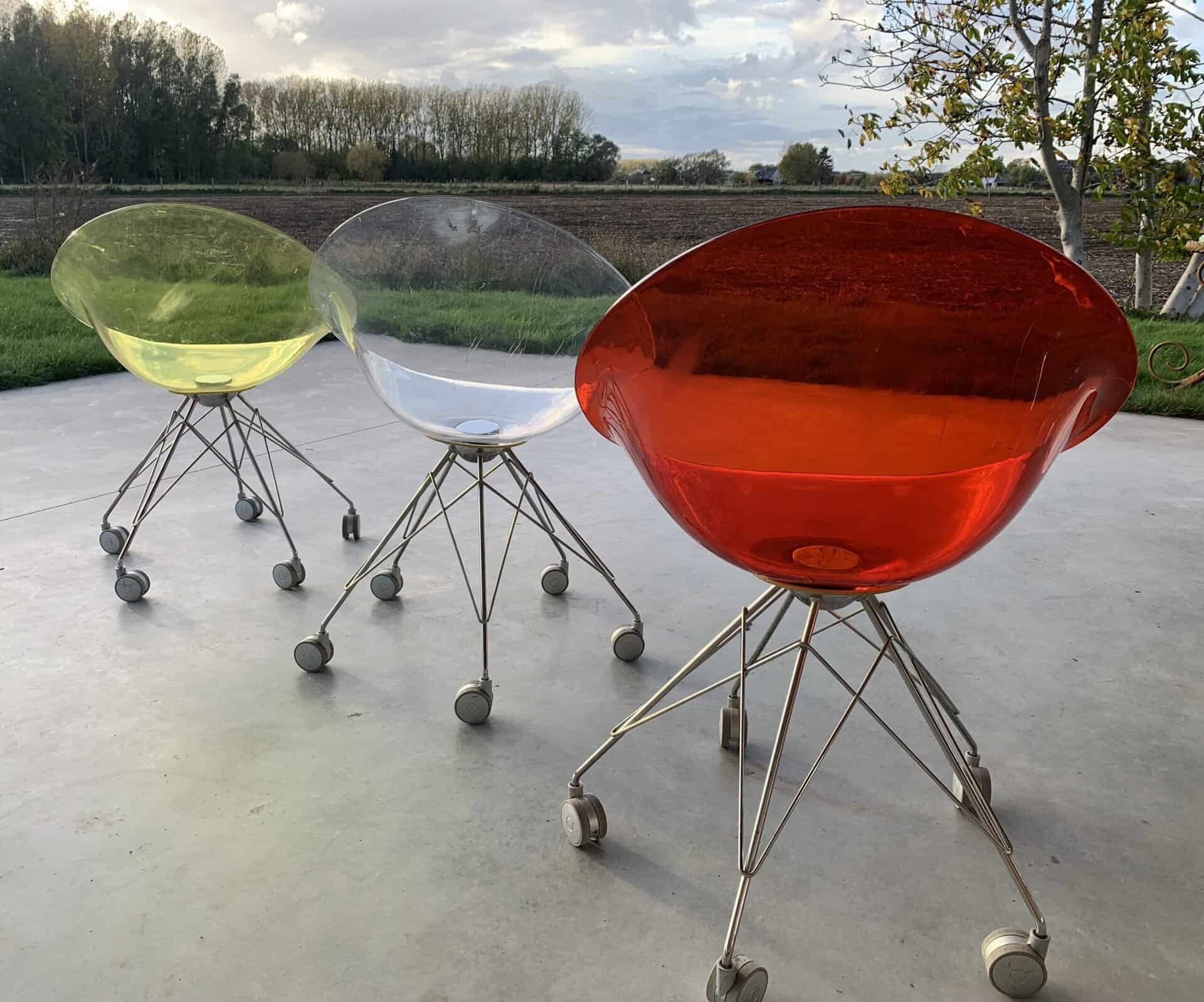 Drie moderne transparante stoelen – geel, transparant en rood – staan op een terras met metalen poten en wielen. Op de achtergrond zijn groen gras, bomen en een bewolkte lucht te zien.