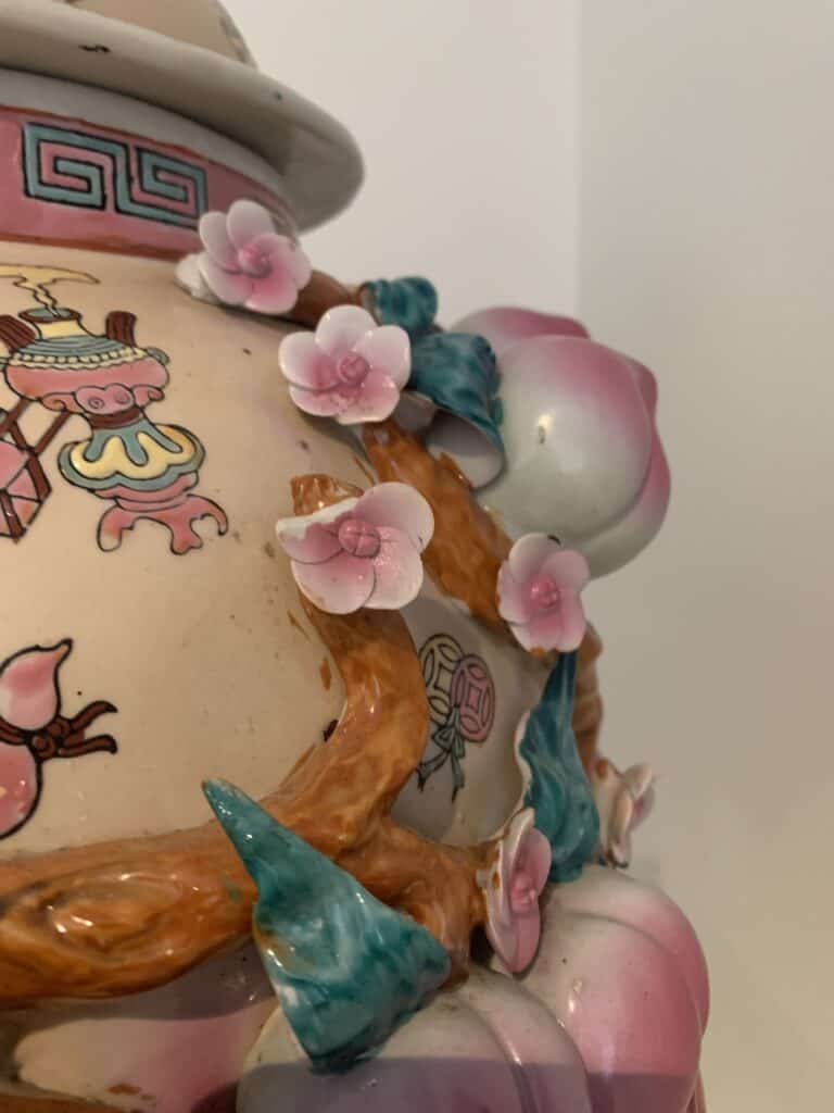 Een close-up van een decoratieve keramische vaas met roze en witte bloemen, groene bladeren en ingewikkelde geschilderde patronen in roze, turquoise en gouden tinten op een crèmekleurige achtergrond.