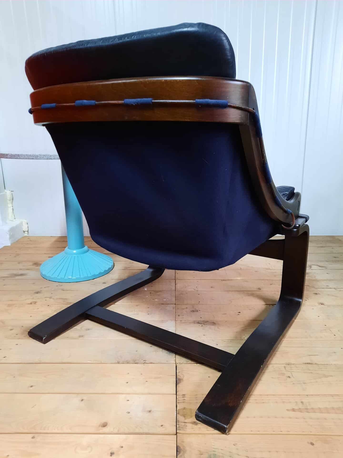 Een loungestoel van donker hout en marineblauwe stof met een gebogen frame en een zachte rugleuning, geplaatst op een houten vloer naast een turquoise tafeltje tegen een witte lambrisering.