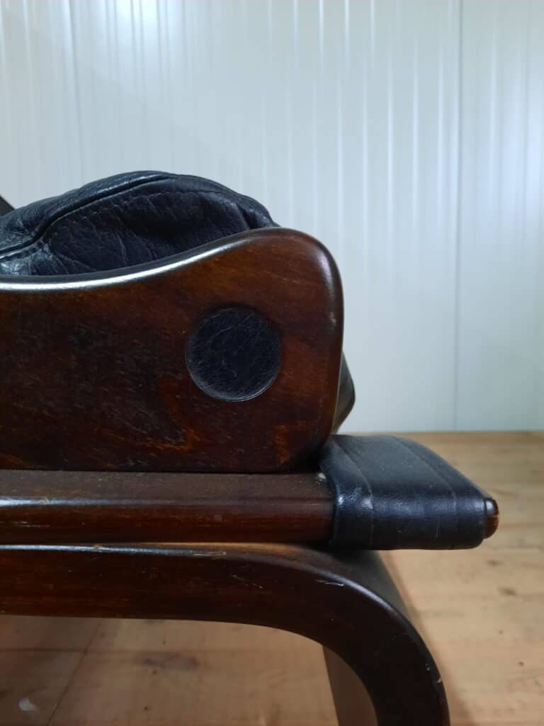 Close-up van de zijkant van een houten stoel met armleuningen van gepolijst donkerbruin hout, zwarte leren bekleding en een lichtgekleurde lambrisering op de achtergrond. De vloer is ook van hout.