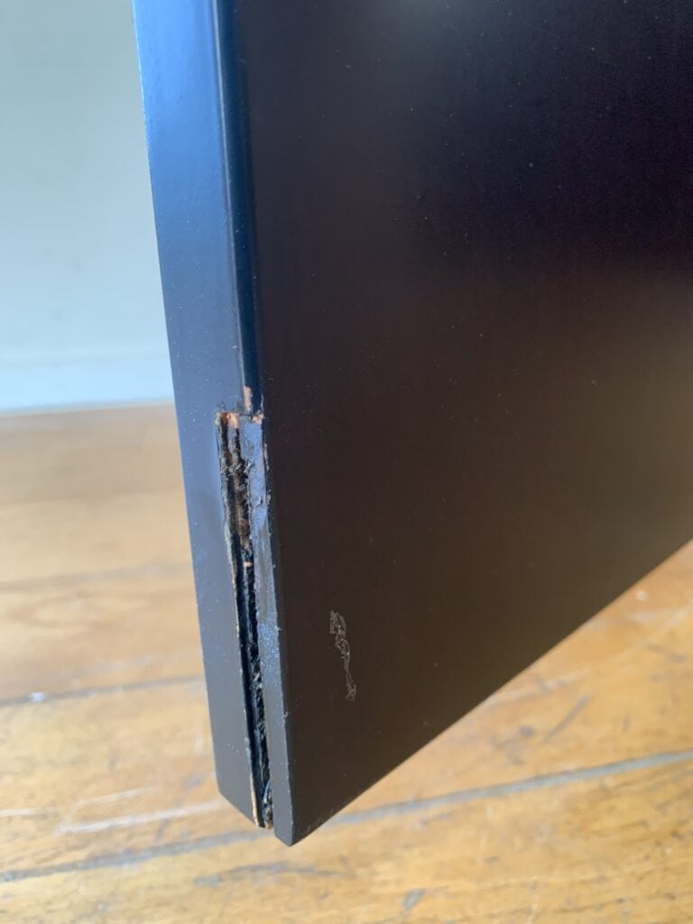 Close-up van een zwart houten paneel met een beschadigde hoek, met afgebladerde verf en zichtbaar, versplinterd hout. Het paneel rust op een houten vloer met zichtbare nerf.