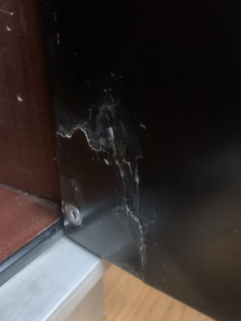 Close-up van een zwart metalen oppervlak met zichtbare krassen, scheuren en slijtplekken vlak bij een schroef, naast een bruin houten paneel en een zilverkleurige metalen rand aan de onderkant.
