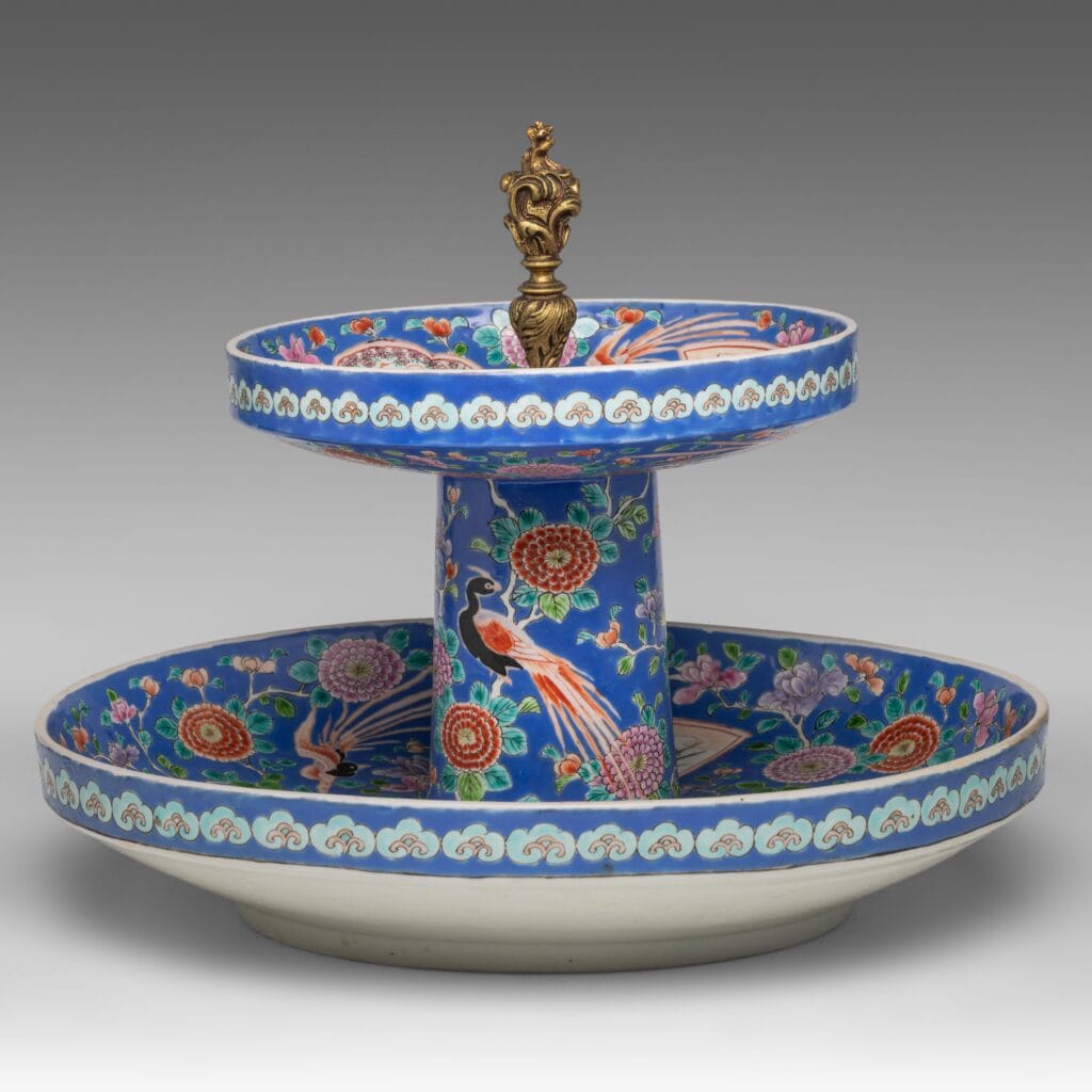 Een decoratieve, tweelaagse porseleinen schaal met blauwe bloemen- en pauwmotieven, geaccentueerd door een gouden sierlijke knop bovenop. De onderste laag is breder en beide hebben ingewikkelde, kleurrijke patronen.