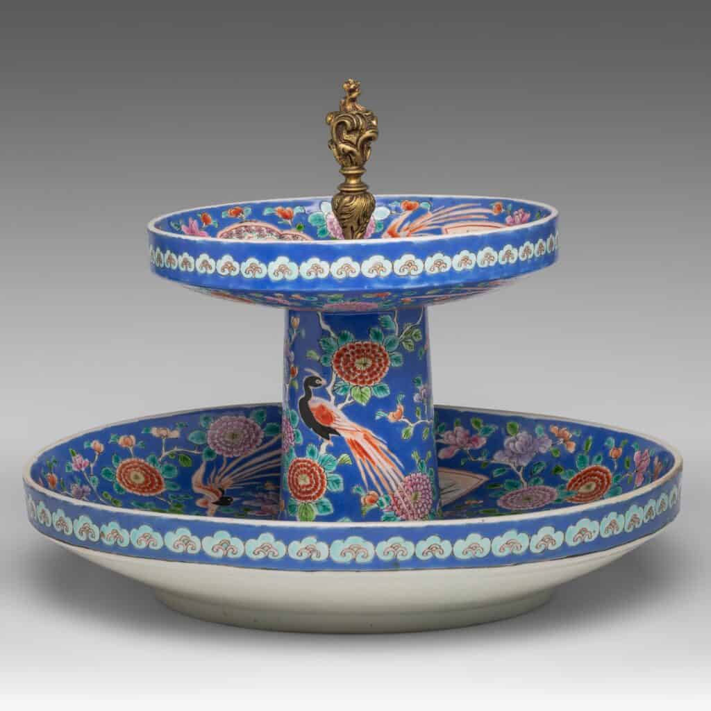 Een porseleinen serveerschaal met twee lagen, met een blauw bloemen- en pauwenpatroon, versierd met gouden details en bekroond met een sierlijke gouden knop.