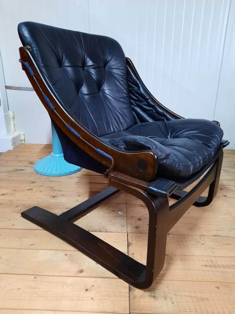 Een zwarte leren loungestoel met gecapitonneerde kussens, een donkerhouten frame en een lage, achteroverleunende vormgeving staat op een lichte houten vloer naast een blauwe tafel met voetstuk en tegen een witte lambrisering.