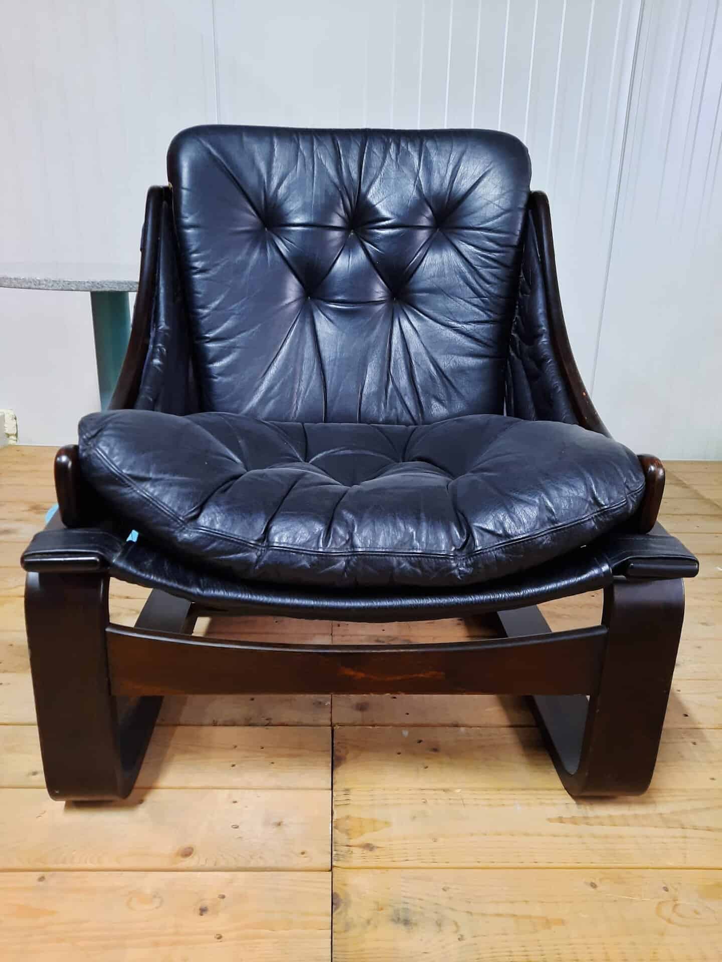 Een zwarte leren fauteuil met een gecapitonneerd kussen en een gebogen houten frame, geplaatst op een houten vloer tegen een lichtgekleurde lambrisering.