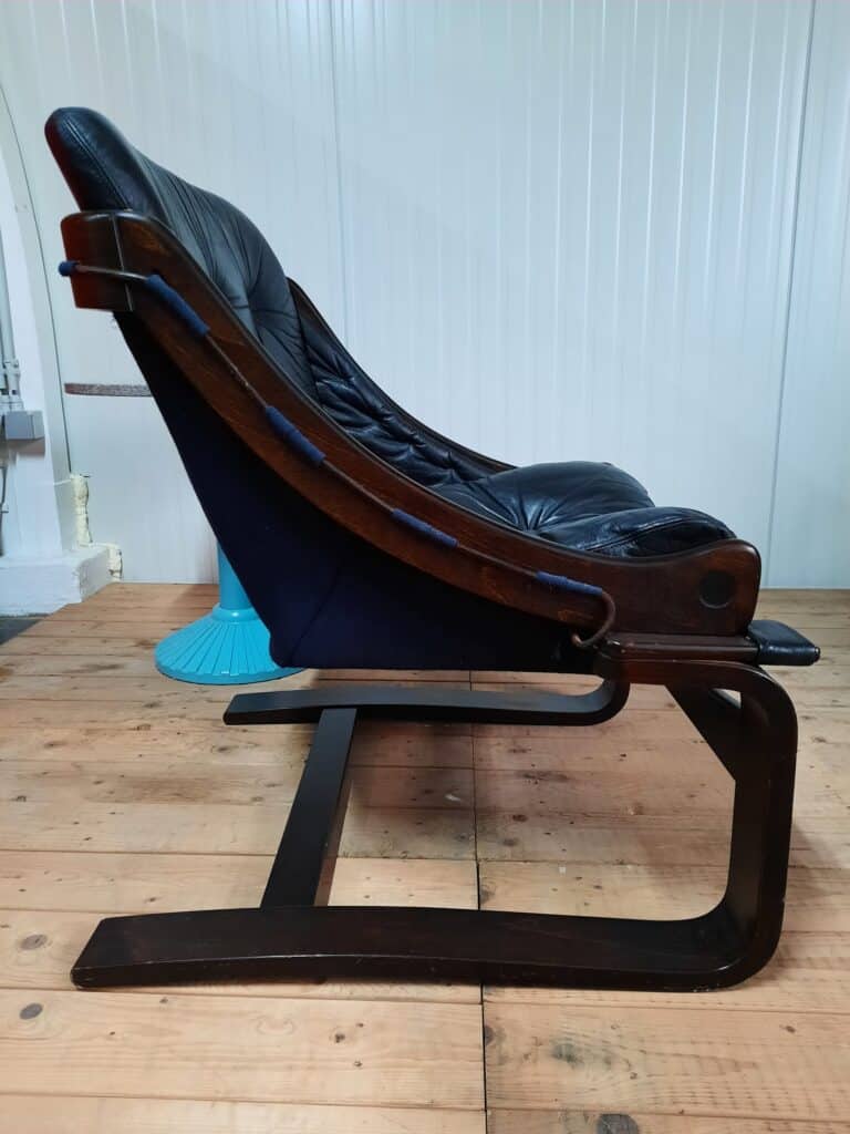 Zijaanzicht van een vintage leren fauteuil met een donker houten frame, geplaatst op een houten vloer tegen een witte lambrisering. Op de achtergrond is een turquoise voetstuk zichtbaar.